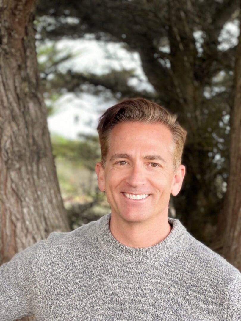 Smiling person wearing gray sweater by trees